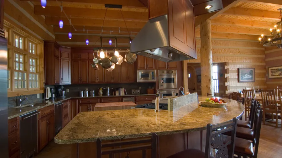 A rustic kitchen with wood log walls, a large granite island, stainless steel appliances, and dark wood cabinets.